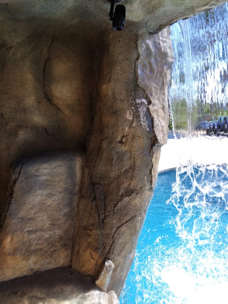 View from inside an artificial rock grotto looking through a curtain waterfall to a bright blue pool