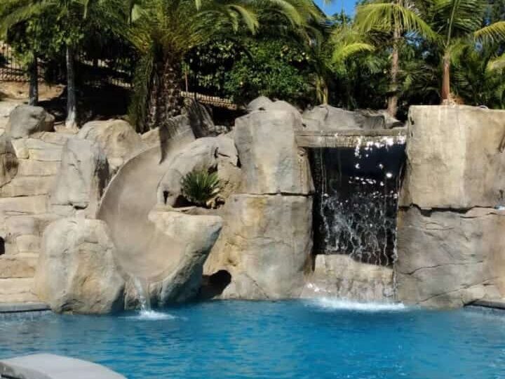 Swimming pool with a sculpted artificial rock slide and grotto waterfall surrounded by palm trees