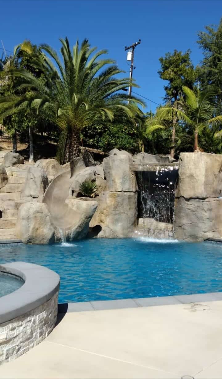 Swimming pool with a sculpted artificial rock slide and grotto waterfall surrounded by palm trees